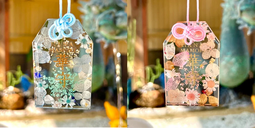 兩個透明護身符懸掛著,左側繫著藍色結,右側繫粉色結,內有花卉裝飾,背景模糊,陽光明媚,顯得寧靜