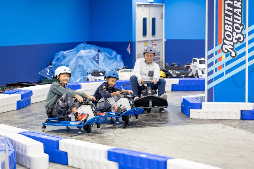 三個戴安全帽的人在室內賽道上駕駛卡丁車,背景為藍色牆面,上有「Mobility Square」文字。氣氛愉快