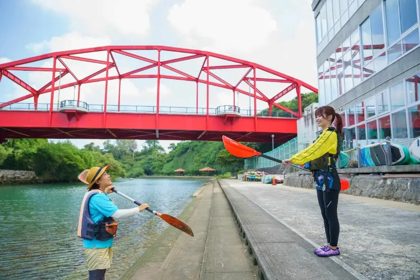兩名女子在河岸邊手持劃槳交流,一人站在岸邊微笑,一人靠近水面;背景是紅色拱橋、綠樹和玻璃建築,氛圍輕鬆明亮