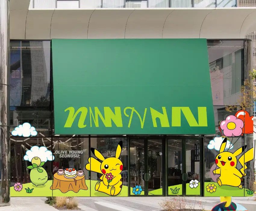 綠色藥妝店外觀，門面貼滿皮卡丘與花朵卡通，前方有蛋糕桌；招牌寫著 OLIVE YOUNG SEONGSU，明亮俏皮。