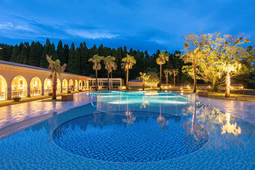 夜幕下的泳池,水面反射藍色光亮,周圍有棕櫚樹和燈光裝飾的建築,環境寧靜優雅,背景為樹林