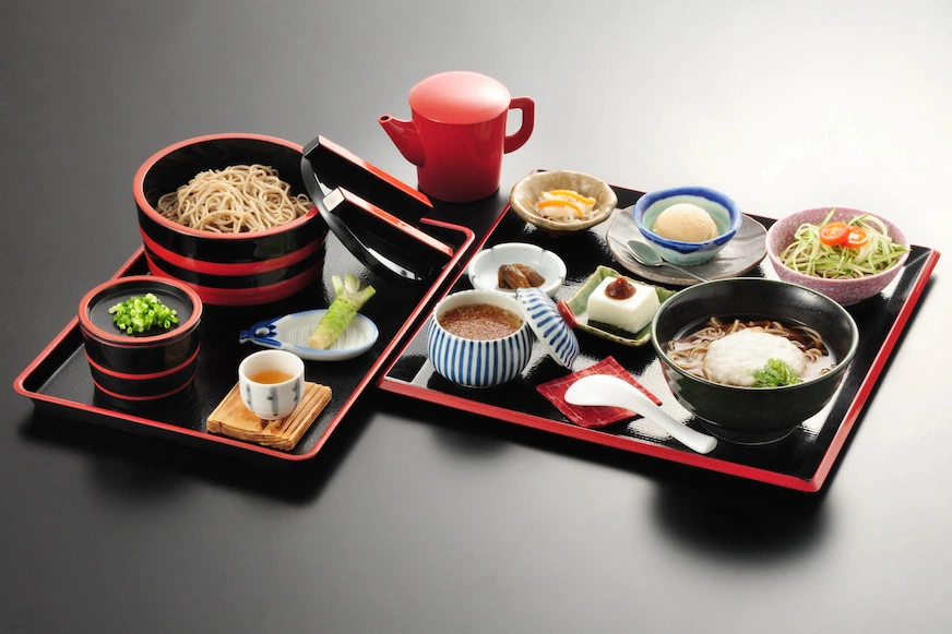 兩份日式餐點擺在黑色托盤上,內含麵條、豆腐、茶杯、調味料等食物。紅色容器和黑色背景形成對比