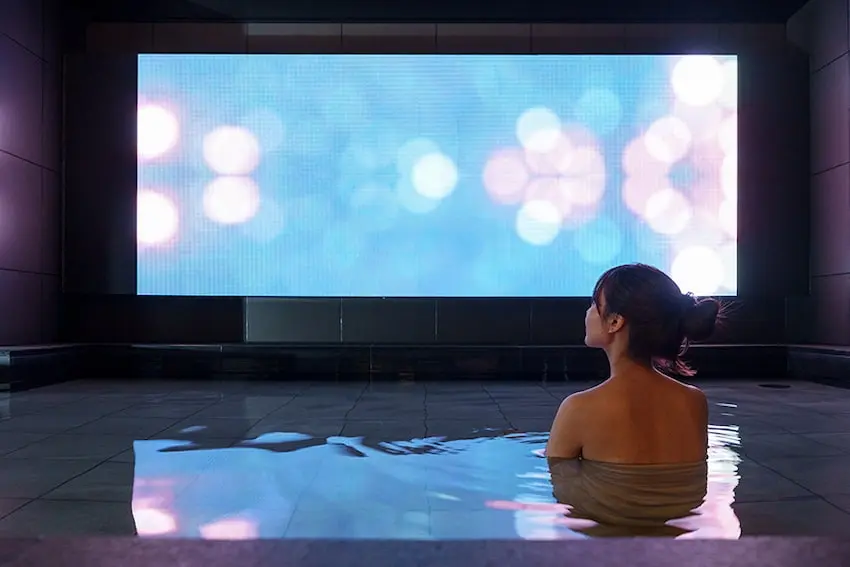 建築內,一位女性坐在水池中,凝視前方巨大的藍色虛光屏幕。環境寧靜,牆壁深色,氛圍放鬆