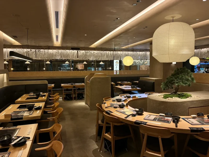 餐廳內部,木製桌椅環繞小盆栽,牆上掛有白燈,環境溫馨。桌上擺放菜單和餐具,燈光柔和