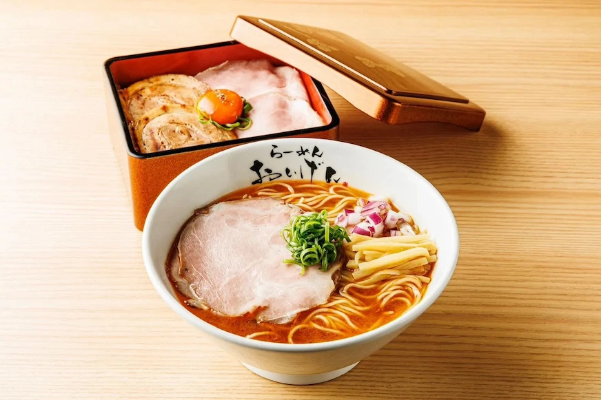 白色碗中盛有拉麵,上有叉燒、蔥花、竹筍。旁邊棕色盒子內放有切片肉和番茄。木質桌面背景