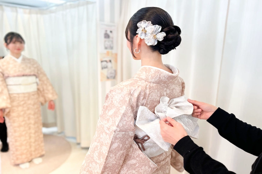一名女子穿著淡粉紅色和服,後置白色花飾。另一人正在調整她的腰帶背景為白色窗簾。氛圍溫馨寧靜