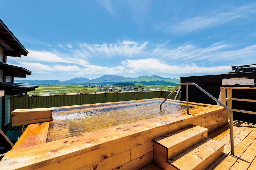 陽光明媚的天空下,一個木製露天溫泉,背景是遠山和綠色田野,氣氛寧靜舒適