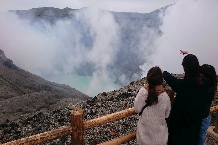 三個人站在木製護欄旁,指向冒著白煙的火山口,背景是灰色岩石和藍綠色湖水,氣氛寧靜