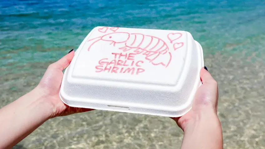 雙手舉著白色外帶盒,盒蓋上畫著粉紅蝦和THE GARLIC SHRIMP字樣,背後是清澈海水與海灘,氛圍輕鬆清爽