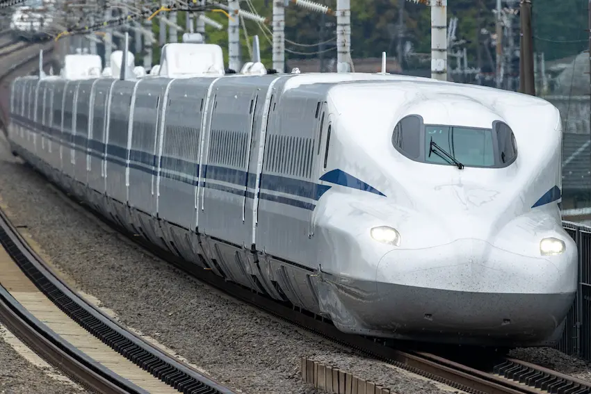 白色新幹線列車快速行駛在軌道上,車身有藍色條紋。背景是電線桿和綠色樹木,展現現代與自然的融合