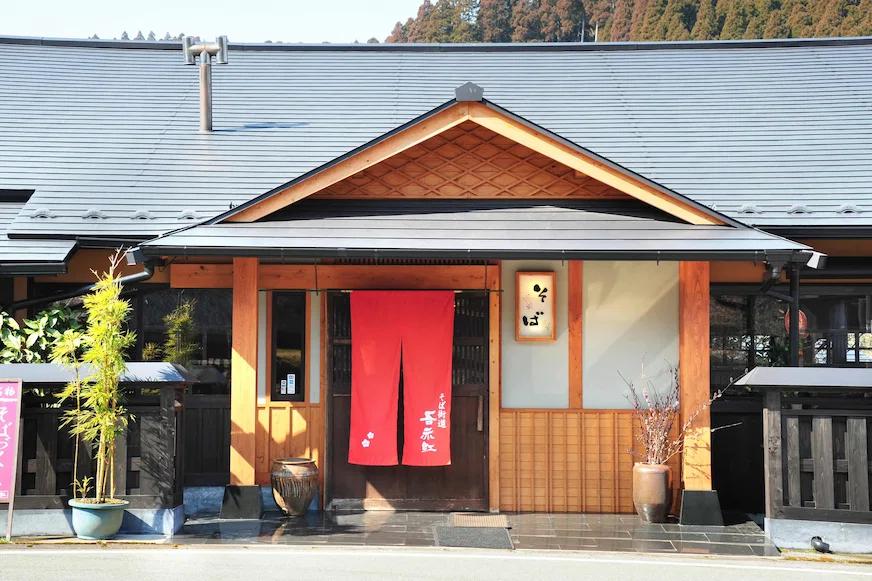 日本傳統房屋外觀,紅色門簾上有白色文字和圖案。屋頂為灰色,背景有竹子和樹木,整體氛圍寧靜