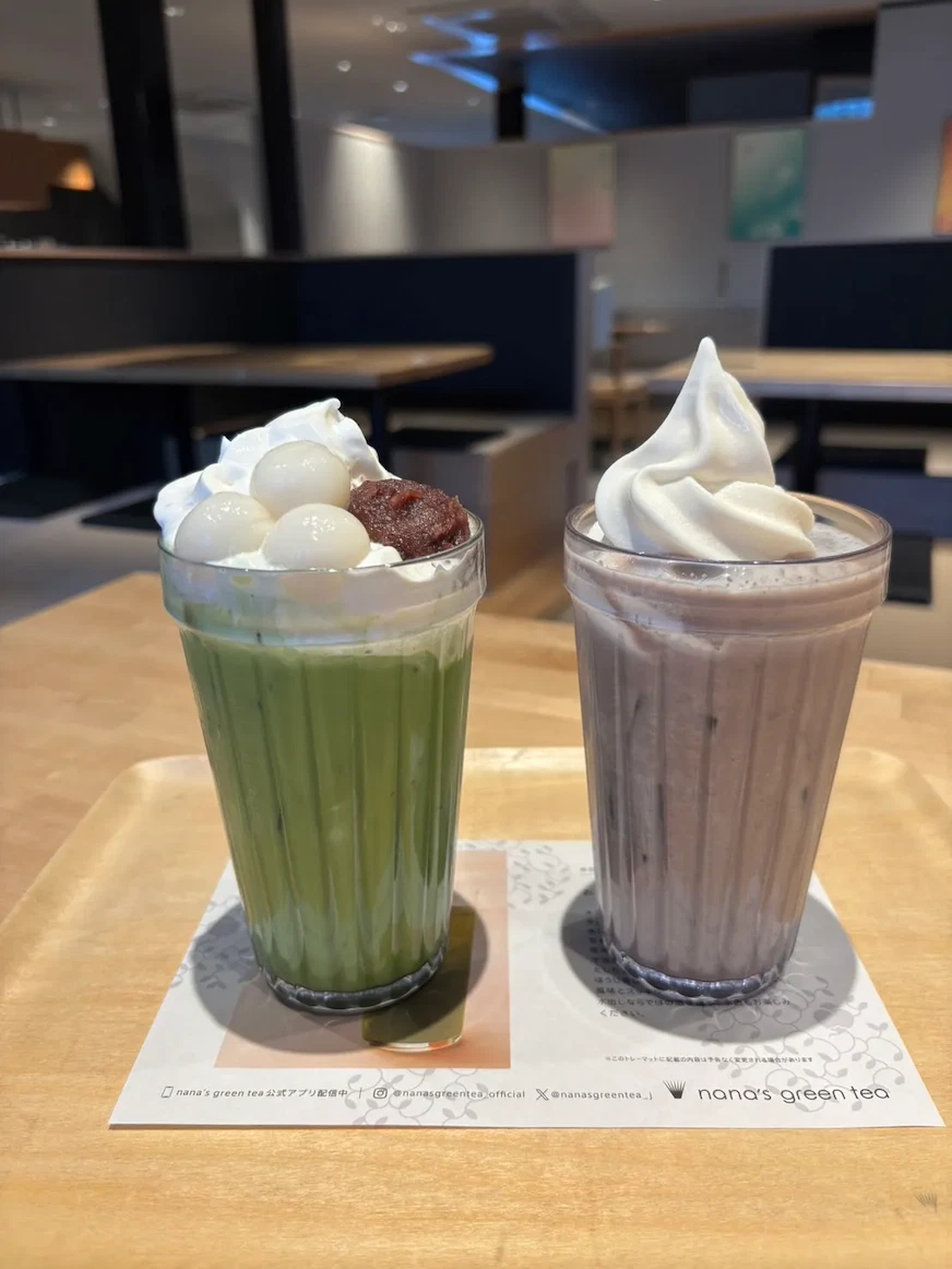 兩杯飲品擺在木托盤上,左邊是抹茶牛奶配白玉和紅豆,右邊是紫色奶油飲品。背景為現代咖啡廳環境