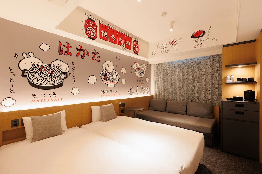 飯店房間,牆上有可愛插畫和日文,展示福岡特色美食。房間內含兩張床、沙發和簡約家具,燈光柔和溫馨