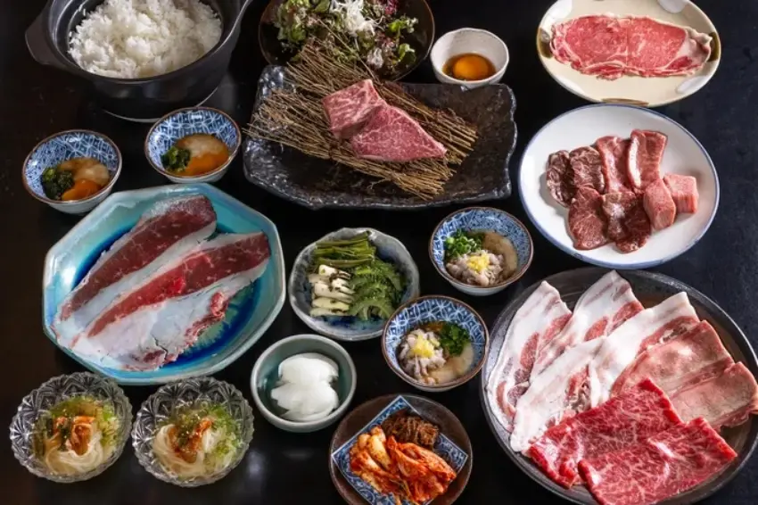 黑色桌上擺滿日式烤肉套餐:多盤生牛肉、五花肉、牛排,配白飯、蔬菜、泡菜和小碟蘸醬,豐盛誘人