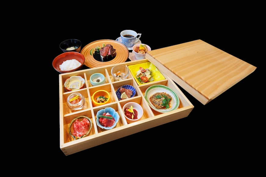 黑色背景上一套餐盒展示多道美食,包括壽司、小菜和甜點,旁有白飯和咖啡,餐具顏色豐富,配色精美