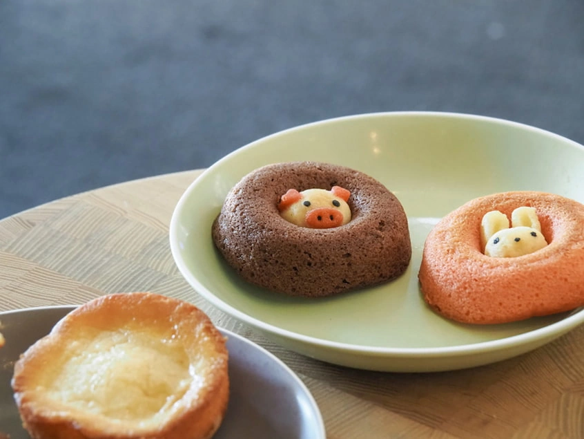 綠色盤中有兩隻可愛動物造型的甜甜圈:一個是棕色豬鼻裝飾,另一個是橙色兔子耳朵。背景為木質桌面
