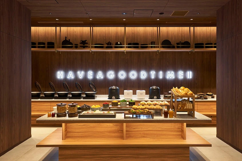 木製自助餐檯上擺滿麵包、果醬及熱水壺,背景牆上有亮燈文字「Have a good time!」。暖色調,現代氛圍