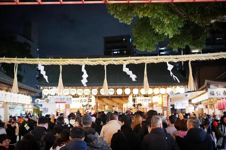 夜晚,人群聚集在傳統燈籠裝飾的神社前,空氣中瀰漫著節日氣氛。背景是城市建築,樹木綠葉在上方