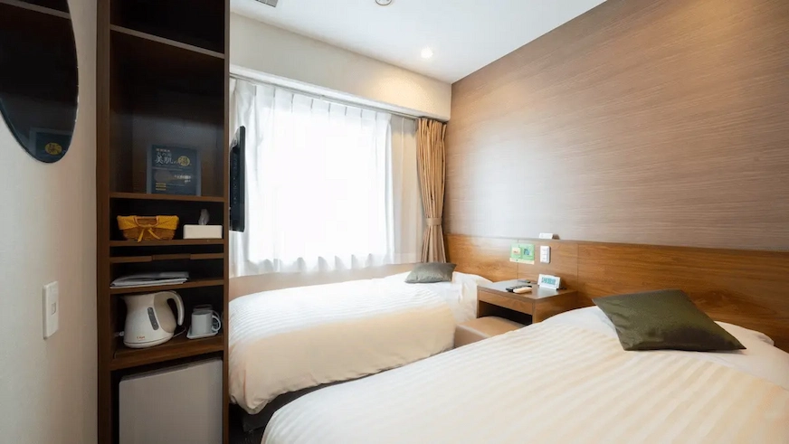 現代雙床飯店房間,木質牆面,兩張白色床單的床上有綠色枕頭。窗外光線充足,房內有書架、電水壺和小冰箱。氣氛舒適整潔