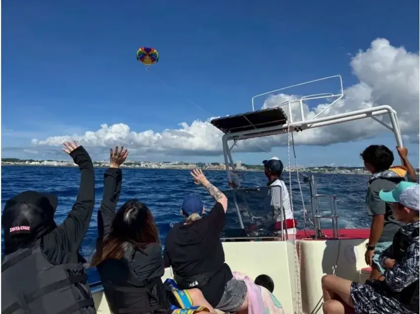 多人坐在遊船甲板上向空中彩色降落傘揮手,藍天白雲下海面遼闊,遠處可見海岸線,氛圍輕鬆歡樂