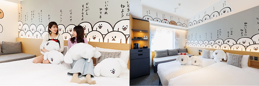 兩名女子坐在白色床上,抱著大白抱枕。背景牆上有卡通圖案和日文。房間明亮,有沙發和裝飾畫,氛圍輕鬆