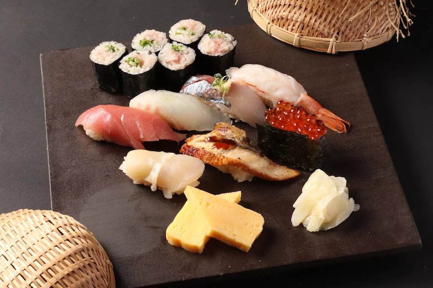 木盤上有壽司拼盤,包括卷壽司和各種刺身,旁邊有笸籮裝飾。黑色背景襯托食物色彩鮮豔