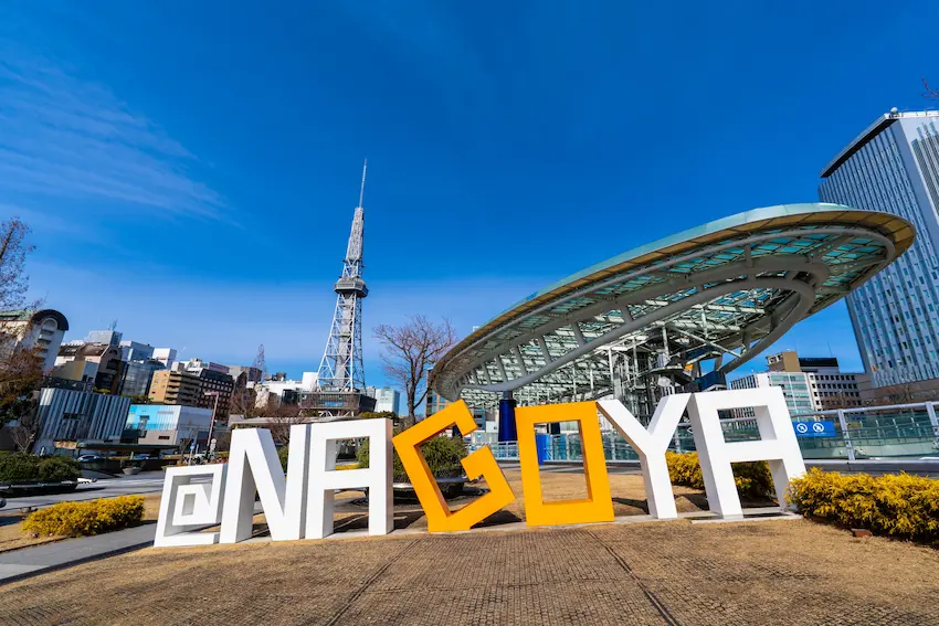 名古屋地標前有巨大的白色和黃色字母,背景是電視塔和現代建築,藍天下顯得清晰明亮,充滿都市活力