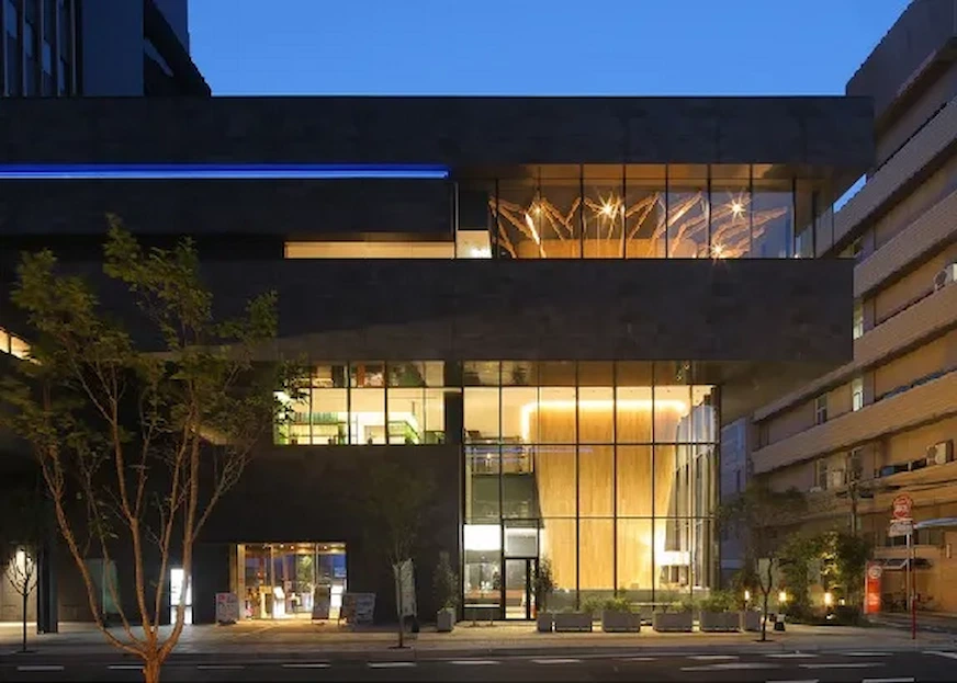 現代建築夜景,玻璃帷幕牆內透出暖黃色光線,藍色天空背景。前景有樹木和城市道路,營造寧靜氛圍