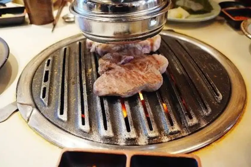 烤肉店裡,幾塊肉在圓形金屬烤盤上炙烤,火焰隱約可見,周圍是餐桌與餐具,氛圍熱鬧