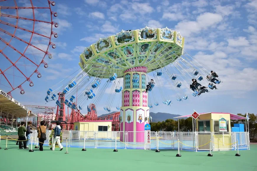 遊樂場內,有人在旋轉的彩色飛椅上,周圍是晴天和摩天輪,前景是幾位遊客