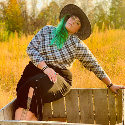 girl with long green hair, plaid shirt, glasses, hat, and tattoo is sitting and leaning back in an a