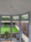A room with curved windows featuring cream blinds, overlooking a green garden with artificial grass and a wooden fence