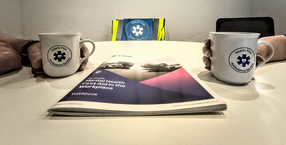 Two mugs with logos are placed on a table beside a workbook titled "Mental Health First Aid in the Workplace." In the background, a jacket with an emblem is visible.