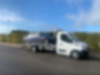 A blue car on the back of a recovery truck on a road next to a hedge.