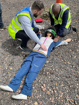 A simulation of a person lying on the ground with individuals attending to them, one is wearing a helmet and the other is wearing a safety vest
