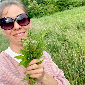 Lächelnde Frau mit Sonnenbrille (Lena Dieterle) hält frisch gesammeltes Mädesüß auf einer Sommerwiese in der Hand.