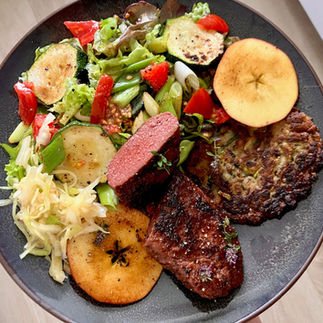 Veganes Gericht mit knusprigem Zucchini-Kartoffelpuffer, veganem Ackerbohnen-Pfeffersteak, gebratenen Apfelscheiben, Blattsalat mit Zucchini, Frühlingszwiebeln und Tomaten sowie einer Portion Krautsalat auf einem dunkelgrauen Teller.