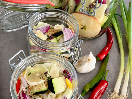 Nahaufnahme von Einmachgläsern mit Zucchini, roten Zwiebeln, Senfkörnern, Knoblauch und essbaren Wildkräutern vor dem Fermentieren, liebevoll geschichtet auf einer Küchenarbeitsplatte.