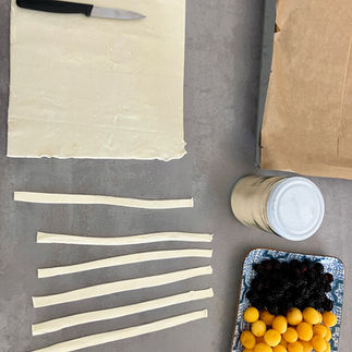 Blätterteig wird mit einem Messer in gleichmäßige Streifen geschnitten, daneben frische Brombeeren, Mirabellen und ein Glas Zucker – Vorbereitung für vegane Törtchen.