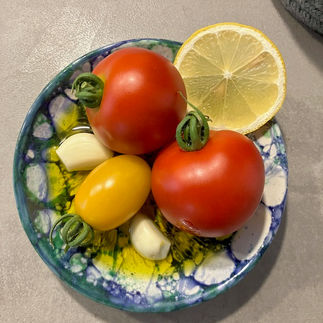 Zwei rote Tomaten, eine gelbe Cocktailtomate, Knoblauchzehen und eine Zitronenscheibe arrangiert in einer bunt gesprenkelten Keramikschale – ein sommerlich-frisches Stillleben.