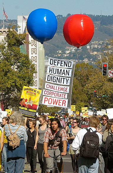 This is Uptown Oakland on the day of a protest demanding a just and sustainable economy. A crowd of diverse races, genders, and ages marches down Broadway. It's mid-day and sunny. In the middle of the image is a tall, vertically-oriented banner held aloft in the air by two huge balloons – one red, the other blue. The sign says “Defend Human Dignity - Challenge Corporate Power.”