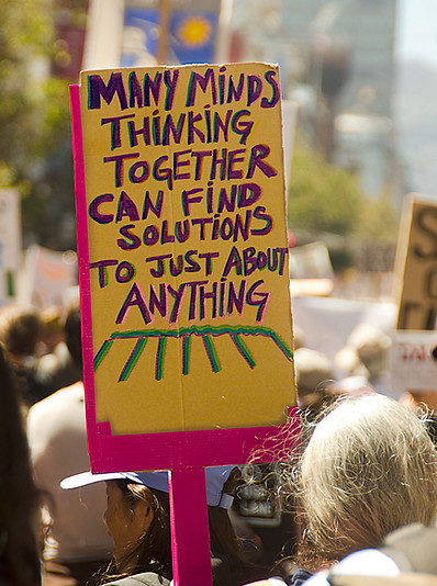At a crowded march for social justice, an activist carries a bright, colorful, hand-painted sign. Its message is “Many Minds Working Together Can Find Solutions to Just About Anything.”