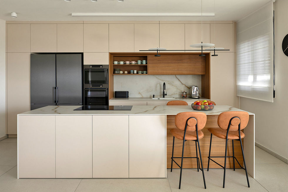 מטבח מודרני ויוקרתי בחלל פתוח, המשלב אי מרכזי במראה שיש בהיר עם ארונות בגוון חול ונישות עץ חמימות.