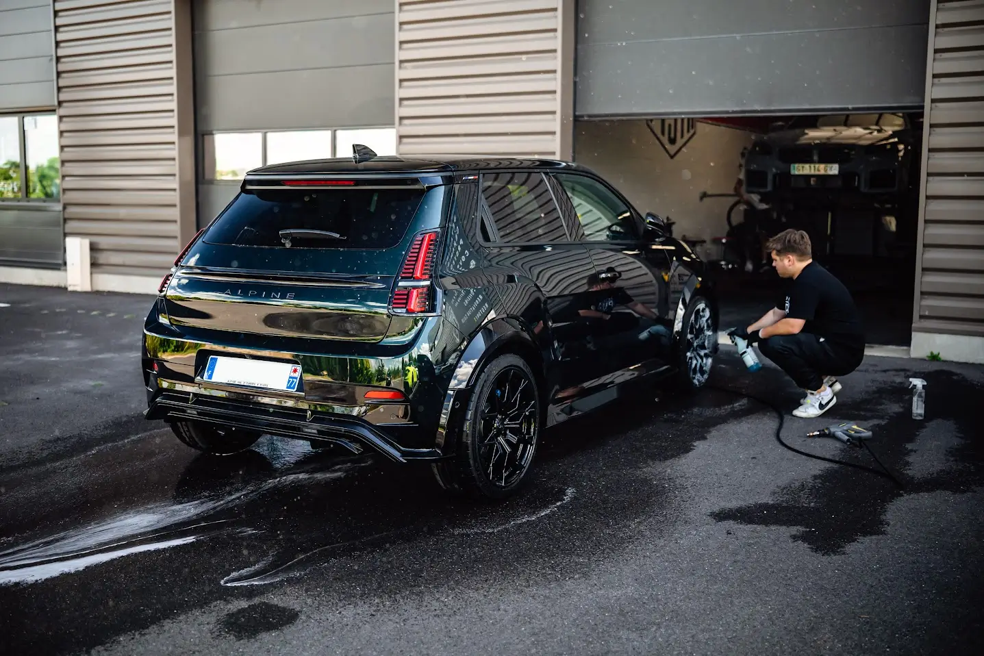 Vue arrière d'une citadine sportive noire brillante en cours de lavage technique à l'extérieur de l'atelier PISTA, un technicien en arrière-plan appliquant un jet d'eau.