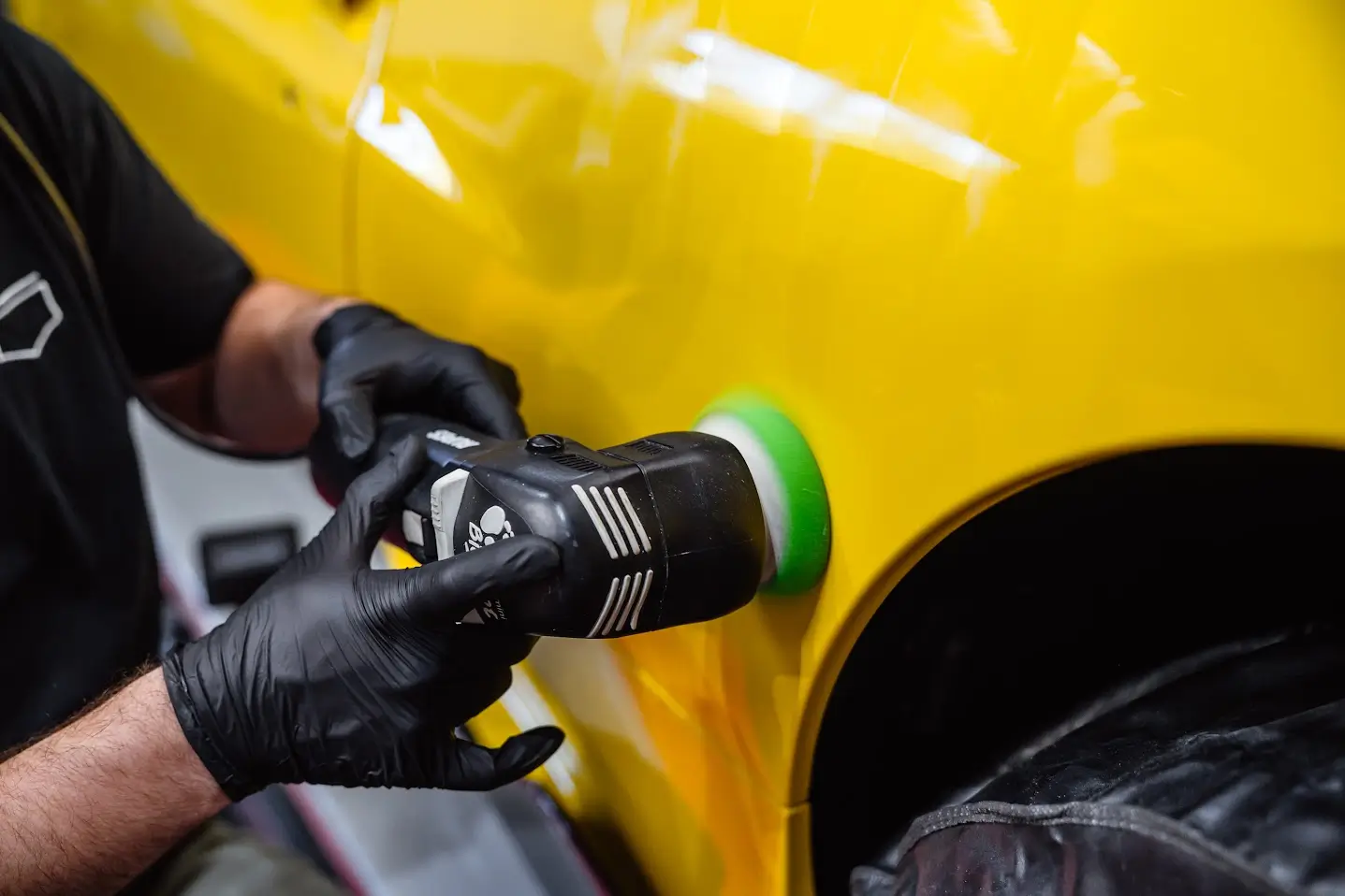 Gros plan sur les mains gantées d'un spécialiste du detailing PISTA maniant une polisseuse orbitale professionnelle avec un tampon vert sur la carrosserie jaune vif d'une voiture de sport.