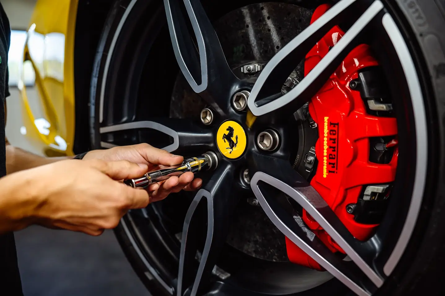 Gros plan sur les mains d'un technicien PISTA utilisant un outil de précision pour le démontage minutieux d'un écrou de roue de Ferrari, révélant le logo central du cheval cabré et l'étrier de frein rouge Ferrari