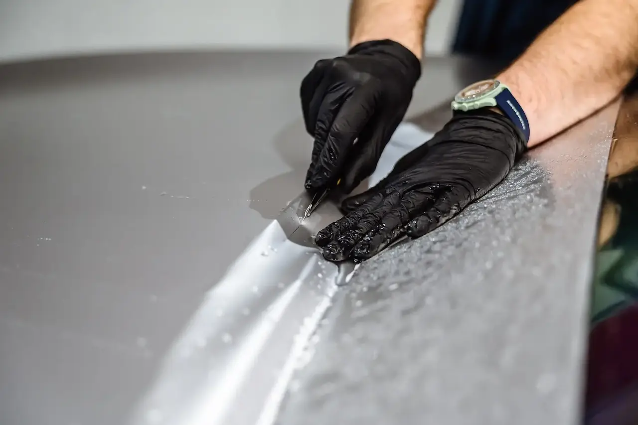 Mains d'un installateur certifié PISTA, portant des gants noirs, effectuant une découpe de précision chirurgicale sur un film de protection carrosserie (PPF) gris mat appliqué sur un capot de voiture.