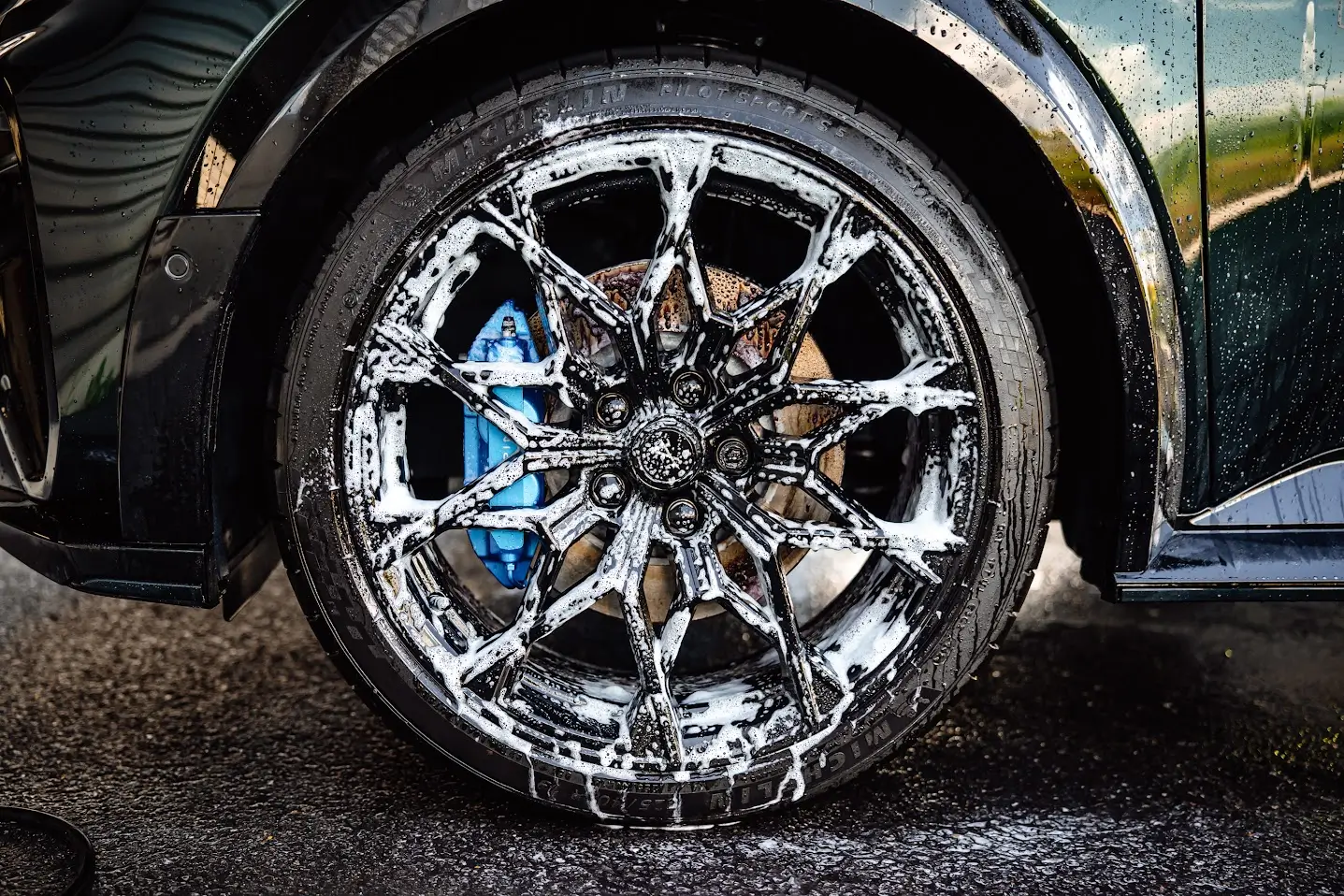Détail d'une jante sport noire multi-branches en plein processus de nettoyage technique, recouverte de mousse de nettoyage active pour le detailing, avec un étrier de frein bleu vif visible.