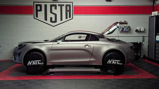 Coupé sport gris sur pont dans un atelier, capot ouvert, panneau PISTA au mur, ambiance technique.