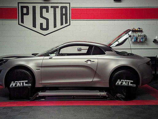 Coupé sport gris sur pont dans un atelier, capot ouvert, panneau PISTA au mur, ambiance technique.
